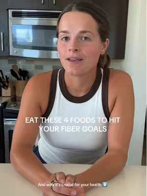 A woman is seated in front of a kitchen. The image is composed of a medium shot, with the woman as the central subject. She is wearing a white tank top with a brown trim and her arms are resting on a surface in front of her. The background includes kitchen cabinets and a stainless steel microwave. Text overlay is present, positioned above them.