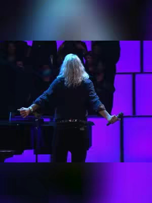 A man with long, white hair stands center stage, arms outstretched, holding a microphone in his right hand. He is seen from the back, dressed in dark clothing. Behind him, a chorus of individuals can be partially seen, with purple lighting illuminating the background, creating a concert or performance setting.