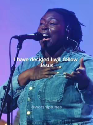 A medium shot captures a Black woman with locs, singing into a microphone with a backdrop of purple and blue lighting. The woman is wearing a denim jacket and has her hand against her chest. Text overlays at the bottom and top, and a microphone stands close by.