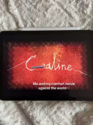 An iPad displaying the title of the movie "Coraline" in a stylized font, set against a fiery red background. The iPad rests on a soft, white, textured surface, possibly a blanket or bedding. A white stylus sits atop the iPad. The text, "Me and my comfort movie against the world" appears below the title.