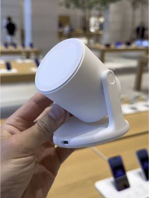 A hand holds a white, cylindrical spotlight device. The spotlight sits on a white stand, with its light-emitting end angled slightly upwards. The background is a bright, modern space that appears to be an Apple store.