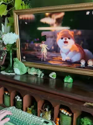 A person is sitting in front of a desk that holds a computer screen showing a game with a cute animated dog on-screen. There are various decorative items around the desk, including a green plush frog and a vase of flowers.