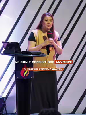 A woman with long brown hair, wearing a yellow vest over a black shirt, stands behind a podium, holding a microphone. The background features diagonal white lines. Text overlays read 'WE DON'T CONSULT GOD ANYMORE' and the social media handle '@LIGHTABLAZEMEYCAUAYAN'.