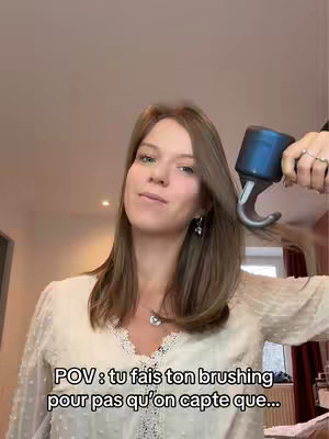 A young woman with long, light brown hair is holding a hair dryer in front of her face giving the impression she is brushing her hair, with a neutral expression on her face. Behind the woman is a ceiling with recessed lighting, some of the room walls, and a window. The woman is wearing a white blouse. Text is added to the image which reads: POV: tu fais ton brushing pour pas qu'on capte que…