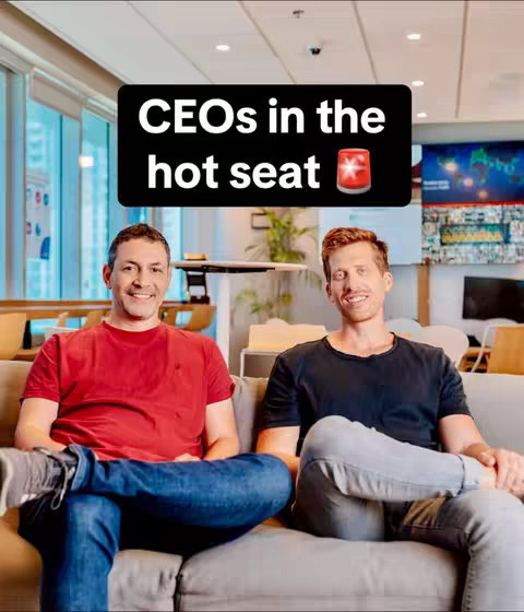 Two men are seated on a light gray sofa in an office setting. One man wears a red t-shirt, while the other is in a black tee. A text overlay reads, 'CEOs in the hot seat'. They sit in front of a large window, with a world map in the background.