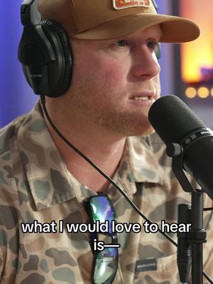 A close-up shot of a man wearing a brown cap, headphones, and a camouflaged shirt. He is holding a microphone and has text overlayed at the bottom of the image.