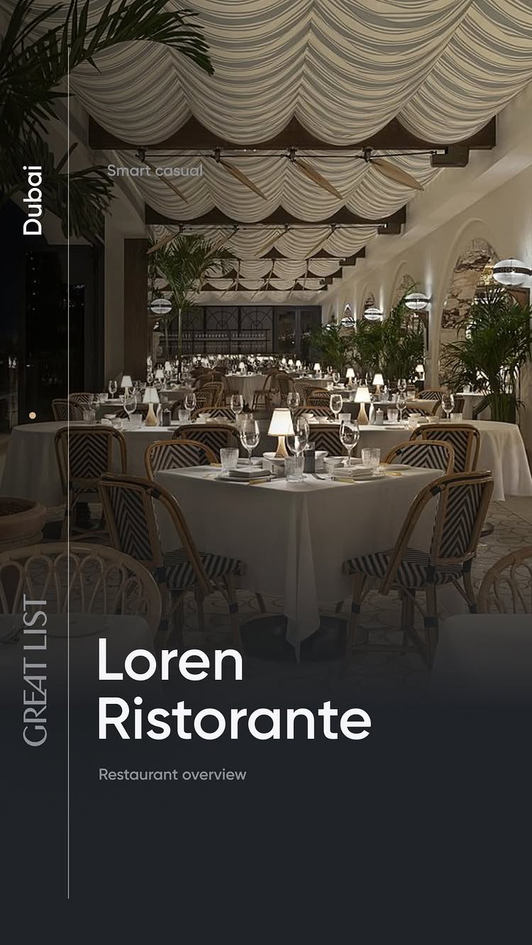 An interior shot of a restaurant at nighttime. The main view shows a large dining area with tables set for dinner. White tablecloths paired with chairs that feature black and white striped upholstery dominate. Overhead, the ceiling has a draped design with lighting. Further in the distance, there are more tables, with plants adding decor. The color palette is primarily dark, with the ambiance suggesting elegance.