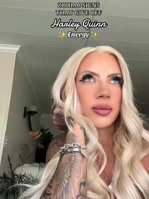 A selfie-style shot features a fair-skinned woman with long, wavy blonde hair. She is looking upwards with large, dramatic eyelashes and full lips. She has a partial tattoo on her arm and is wearing a bracelet. The background shows a glimpse of the surrounding room with some greenery.