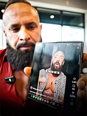 A man with a large beard holds up a phone displaying a video of himself. The phone screen shows a shirtless man with a similar beard and a text overlay. The background suggests an interior space, with bright lighting.