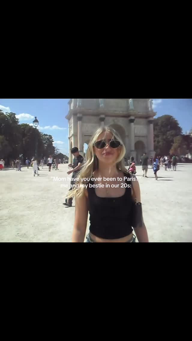 A woman stands in front of the Arc de Triomphe in Paris. She is wearing sunglasses and a black top. Text overlayed reads, \"Mom have you ever been to Paris? me and my bestie in our 20s:\". Many people are in the background.