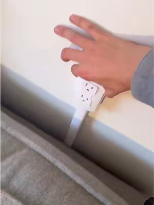A hand holding a white power strip is placed near a white wall and a cushioned surface. The hand is in the frame, and the power strip is hanging down, connected to the wall.
