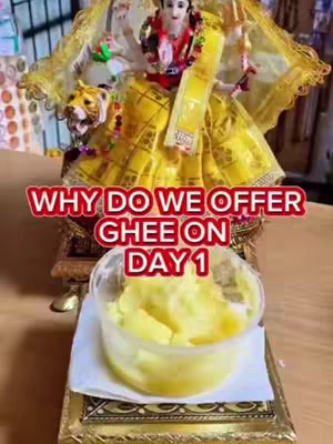 The cover image features a divine statue of a Hindu deity, draped in a golden yellow garment. Positioned in front of the statue is a transparent container holding a generous portion of ghee, a clarified butter, presented on a decorative golden base. The background slightly blurred, suggests the backdrop is indoors. The top of the image features a red banner styled text reading 'WHY DO WE OFFER GHEE ON DAY 1' with a white outline.
