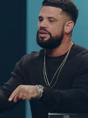 A medium shot captures a man with a neatly trimmed beard and short, styled hair, set against a backdrop of muted teal and dark blue. He wears a black long-sleeve shirt and a silver necklace. His left hand, adorned with a silver watch and bracelet, is extended, pointing forward. The lighting is even, highlighting his facial features. He appears to be speaking animatedly.