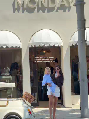 A woman in sunglasses holds a young child, standing in front of a storefront with the word "MONDAY" on top. A vintage car is parked on the left. Light and airy aesthetic.