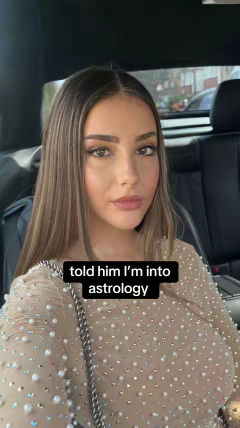 A woman is sitting in a car, facing the camera. She has long, light brown hair, and is wearing an embellished, nude-colored dress. A text overlay says, "told him I'm into astrology". The background shows a blurred view of the outside world.