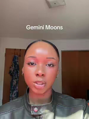 A selfie-style image of a Black woman, medium shot, with soft lighting. She has dark, well-defined eyebrows and is wearing makeup. Her skin is smooth with a slight glow, particularly under bright lighting. Her hair is styled off of her face. The background shows a portion of a room, including a door and a wardrobe.