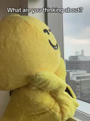 A person in a yellow mascot costume looks pensively out a window. The mascot is large and rounded, with the head featuring a smiling face and large eyes. Outside the window, a cityscape is visible with tall buildings. The image is framed from the mascot's perspective.. Above the mascot is text in white.