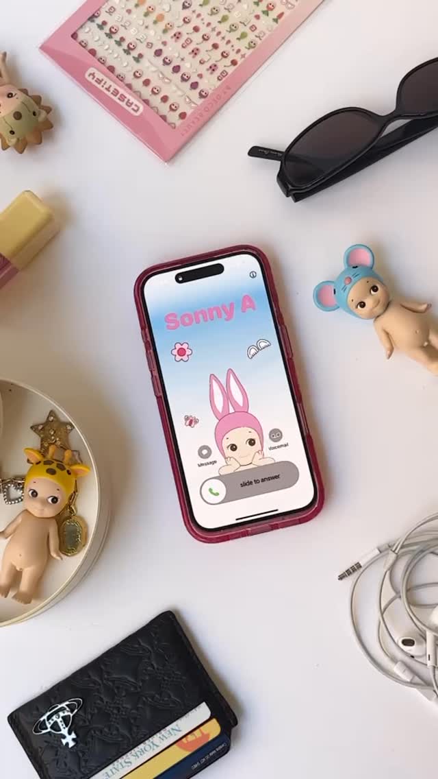 A top-down shot presents various objects on a white surface, including a smartphone displaying a colorful illustration, stationery, sunglasses, and small figurines. The phone screen features the name 'Sonny A' in a playful font above a cartoon character wearing a pink bunny hat. Surrounding the phone are accessories like sunglasses and a black wallet, along with a selection of decorative items.