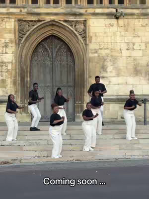 A group of eight people, seemingly a choir, stand on steps in front of a large, ornate building with a large arched doorway. They are dressed in matching black tops and white pants. The text overlay "Coming soon ..." appears at the bottom of the image.