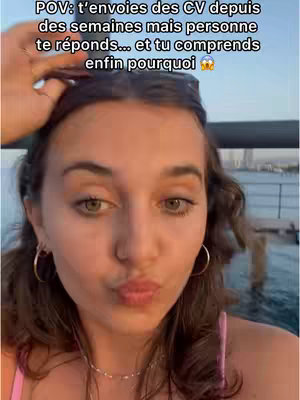A woman with brown hair, holding a strand of her hair back with one hand, while slightly pursing her lips. The background is blurred and appears to be an outdoor setting with water visible.