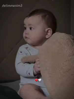 A close-up shot of a baby seated, holding a plush toy. The baby is facing left with a curious expression. The background is a muted shade of brown and tan.