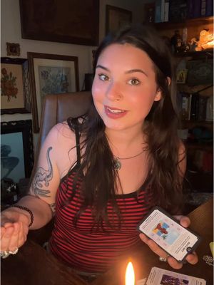 A woman is seated at a wooden table, holding a tarot card and appearing to be engaged in a reading. She is centered in the frame, with a softly lit candle to her right and soft lighting overall. Artwork is displayed on the wall visible behind the woman's head, and a bookshelf is to the right, implying an intimate and personal setting. The woman is wearing a red and black striped tank top and has a tattoo of a dinosaur on her left arm.