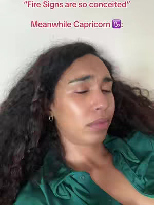 A face close-up shows a person with long, wavy dark hair and a green top. The person's eyes are closed and they have a neutral expression. Overlayed text at the top uses a pink font, while the bottom text indicates a Capricorn zodiac symbol.