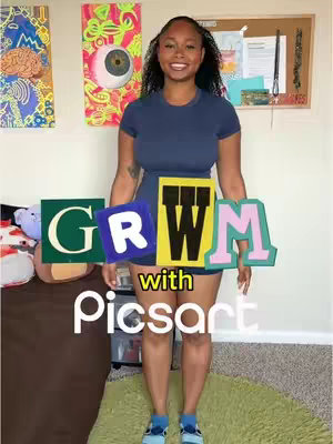 A woman stands in front of a light wall with artistic decorations. The background features abstract art and a corkboard. The foreground displays the text "GRWM with Picsart" in colorful, blocky letters. The woman is smiling and wearing a navy blue dress with blue shoes.