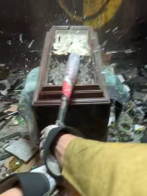 A first-person perspective shows someone breaking a clock with a baseball bat. The image is slightly out of focus. A wooden clock is in the center, with glass being shattered by the bat. The person's arm, wearing a glove, is visible, holding the bat. Other items such as glass and a yellow surface are in the environment. The overall composition is chaotic, suggesting a destructive act.