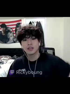 A medium shot shows a young man with dark, curly hair in front of a camera with an American flag on the wall behind him. The man is wearing a black t-shirt and earbuds and is looking toward the camera.  A Twitch logo with his username is overlaid near the bottom of the frame.