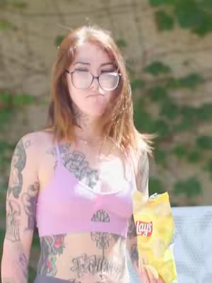 A woman with tattoos is holding a bag of chips against a blurry green background. She is wearing glasses and a pink sports bra.