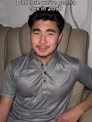 A young man with dark hair and a gray polo shirt is seated, likely indoors, angled to the camera. The background reveals beige-colored seating indicative of an airplane or similar transport. The man's expression is confident, with a slight smile. The composition is a medium shot.