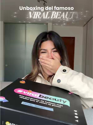 A woman with dark hair and a neutral beige top is holding a large, black box with the words '#viralbeauty' prominently displayed. She's covering her mouth with her right hand in a surprised or excited gesture. The image is brightly lit, and the background is a blurred interior setting, likely a home. Text overlays at the top mention 'Unboxing' and 'Viral Beauty'.