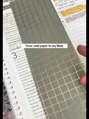 The image shows an open Bible with handwritten notes and a piece of patterned paper being added. The paper is light olive green with a repeating gold design. The Bible pages are visible, with text and some highlighted passages. A hand is seen holding and positioning the decorative paper.