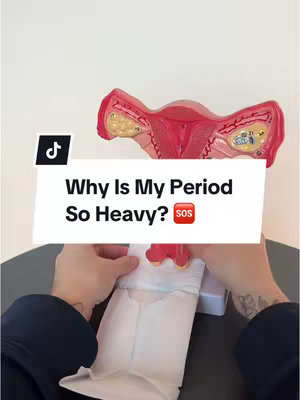 The image shows a close-up view of a model of female anatomy on what appears to be a table or other surface. The model is primarily red and pink, representing the uterus and surrounding structures. Below the model, white material (possibly a sanitary pad or towel) is being held by a person. The background is plain white. A text overlay reads 'Why Is My Period So Heavy?', with a small 'SOS' icon next to it.