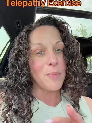 A woman with curly brown hair and a light-colored top sits in a car. The video is framed from inside the vehicle, with the windshield and adjacent window visible. The woman is looking directly at the camera and speaking with an engaged expression. The background is blurred, with natural light suggesting an outdoor setting.