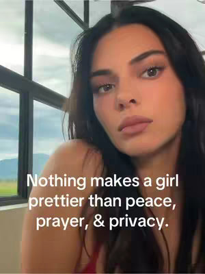 A selfie-style shot featuring a woman with dark hair and a neutral expression. She is facing the camera closely, with soft lighting subtly highlighting her features. Text overlayed states 'Nothing makes a girl prettier than peace, prayer, & privacy.' A window frame is visible in the background.