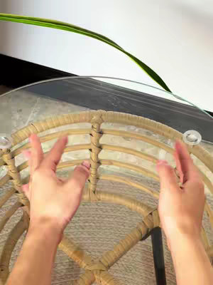 A top-down shot features a person's hands resting on a woven wicker chair. A circular glass table is positioned above the chair, and in the upper-left corner, a green and yellow leaf gracefully curves into the space.