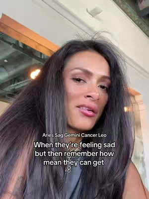 A woman with dark hair and a neutral expression looks directly into the camera. Overlayed text includes the zodiac signs Aries, Sag, Gemini, Cancer, and Leo, with a statement about their emotional tendencies. The background has warm-toned wooden elements, and the overall lighting is natural.