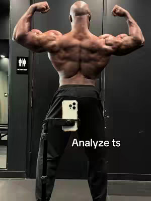 A man with a bald head flexes his back muscles in a weight room, viewed from behind. He's wearing dark sweatpants and the camera, mounted to a tripod, is visible. The image focuses on his physique and is framed to incorporate the cell phone, highlighting both the muscular definition and the media creation aspect.