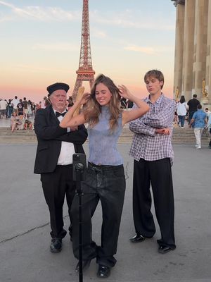 A photo taken in Paris, France, featuring three people in front of the Eiffel Tower. A woman is in the center, holding her hair up, a tripod for a phone is in front of her. There is an older man on the left wearing a suit, bow tie, and a black beret and a younger man to the right wearing a plaid shirt. Other tourists are in the background.