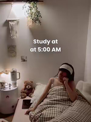 An indoor shot shows a person in bed, appearing to wake up. Text overlayed on the image reads 'Study at at 5:00 AM'. The person is under a brown-and-white checkered blanket, in a white tank top and white headband. Behind them are a cream wall decorated with a small macrame and a plant.