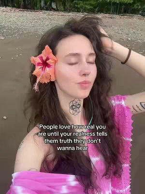 A woman with long, wavy brown hair stands on what appears to be a beach, facing the camera with her eyes closed. She wears a pink and white tie-dye style top and has a large orange and red flower tucked behind her ear. A tattoo on her neck showcases a geometric design, and another tattoo is visible on her upper arm. Text overlayed on the image reads, "People love how real you are until your realness tells them truth they don't wanna hear."