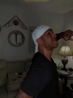 A man is shown in a room, angled slightly down and to the right. He wears a white head covering and a black t-shirt, his face turned up and to the side.