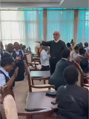 A video still shows Benny Hinn, a pastor with white hair, in a room with many other people. Hinn is gesturing with his right arm outstretched, appearing to be addressing a gathering. Members of the staff are standing and praying. The background shows an interior with large windows.