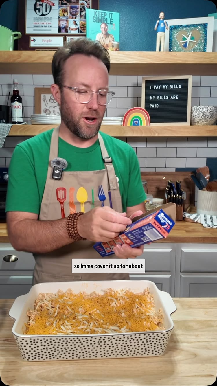 A man in a green shirt and apron is holding a package and looking towards the camera, a casserole dish with a chicken pasta bake in the foreground. The background shows a kitchen setup with shelves and framed artwork.
