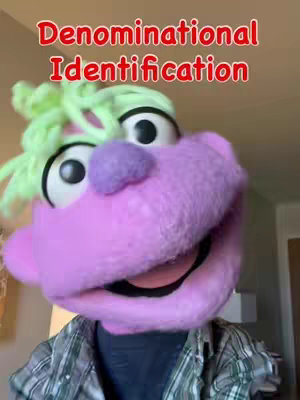A close-up shot of a puppet with a purple face, large eyes, a purple nose, and bright green hair. The puppet is smiling with its mouth open, revealing a peek of the inside of its mouth. The top of its head has green hair. The puppet is set against a background that fades from a tan wall into a room beyond, and the individual operating the puppet can be seen in the bottom through the shirt. Above the puppet's head is text.