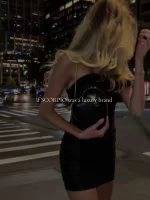 A nighttime shot of a woman walking on a city street. She is wearing a black outfit and carrying a black handbag. Text overlay at the bottom states 'if SCORPIO was a luxury brand'. The background includes city lights and buildings.