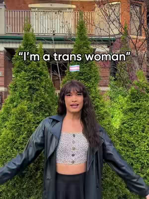 A person stands in front of a lush green backdrop of trees. The person is wearing a black leather jacket over a white, button-down top and black skirt. The background includes a traditional brick building and the text overlay says, "I'm a trans woman." A trans pride flag is also in the background between the trees.