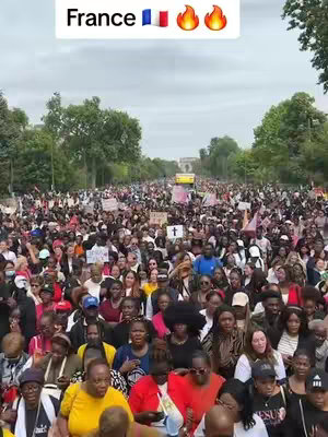 A wide shot captures a massive crowd of people filling a wide street, likely during a march or demonstration. The crowd is dense, composed of individuals of various ethnicities and ages, with many holding signs. Buildings line the sides of the street, and natural light illuminates the scene from above. A text overlay at the top indicates that the location is France.